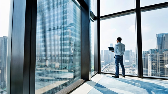 A man standing with a laptop in his hand in a high-rise building with floor-to-ceiling window and lookin at other skyscrapers