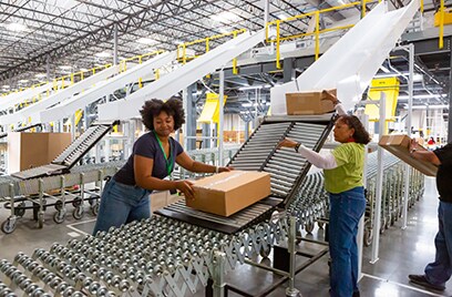 Two women working at a factory where one woman is supervising and the other woman moving pacakges