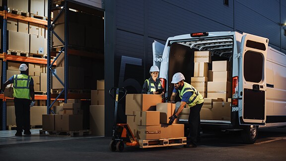 Two workers wearing safety jackets unloading packages from a van in front of a warehouse and another worker standing inside the warehouse