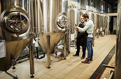 Two male workers talking to each other while standing standing next to a brewing machine